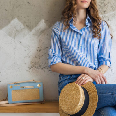 Young attractive stylish woman sitting at wall background, holding straw hat, handbag, wearing blue cotton shirt, jeans, casual outfit, hipster vintage style, spring summer fashion trend, accessories