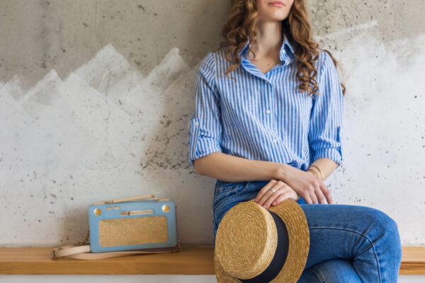 Young attractive stylish woman sitting at wall background, holding straw hat, handbag, wearing blue cotton shirt, jeans, casual outfit, hipster vintage style, spring summer fashion trend, accessories