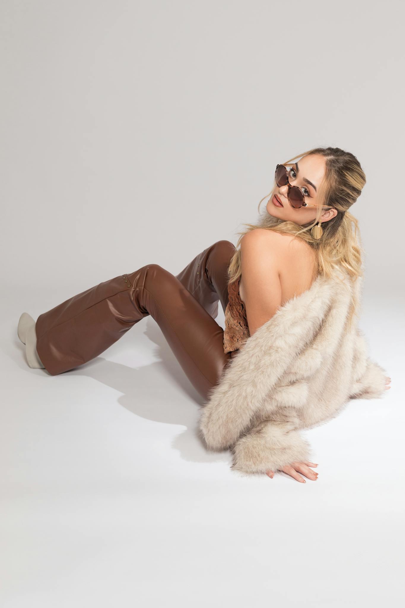 Chic woman wearing sunglasses, furry coat, and brown leather pants on a white background.