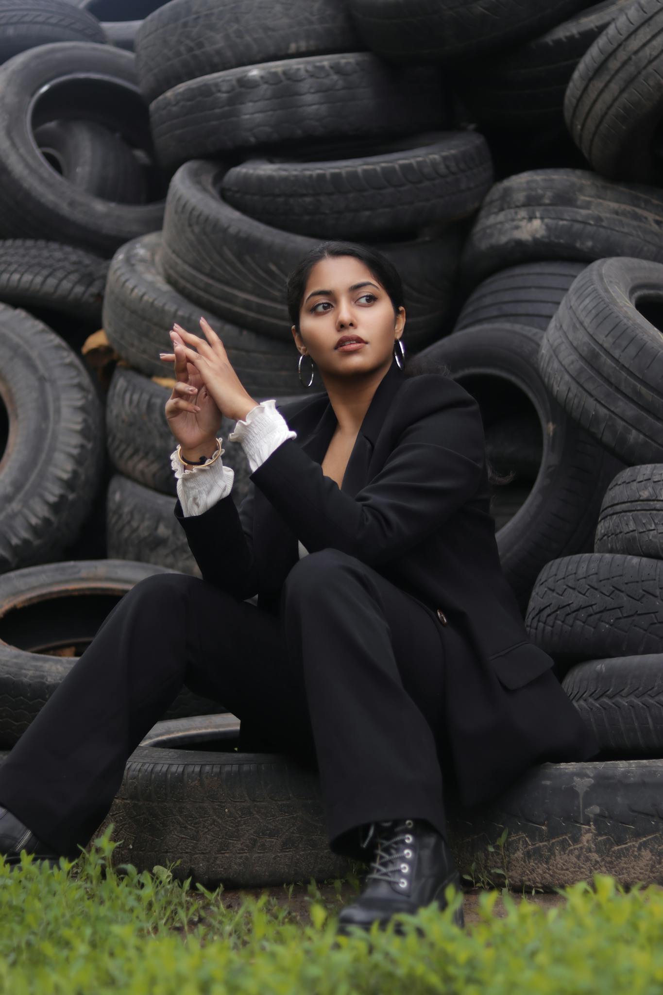 Woman in black blazer sitting against a backdrop of stacked tires, looking contemplative.